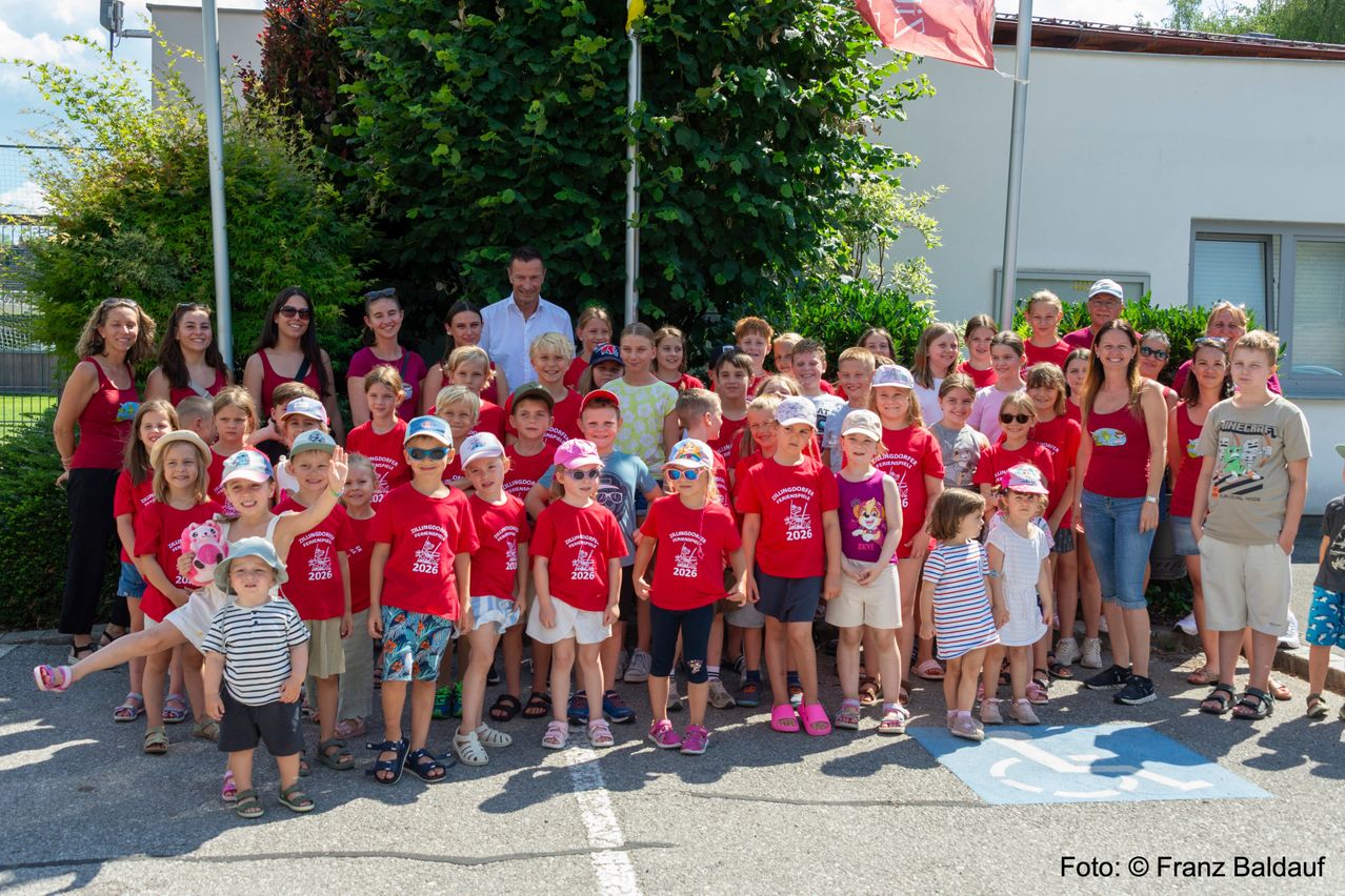 Eine große Gruppe von Kindern in roten T-Shirts mit dem Jahr 2026 darauf, stehen vor einem Gebäude. Die Erwachsenen und Kinder posieren für ein Foto.