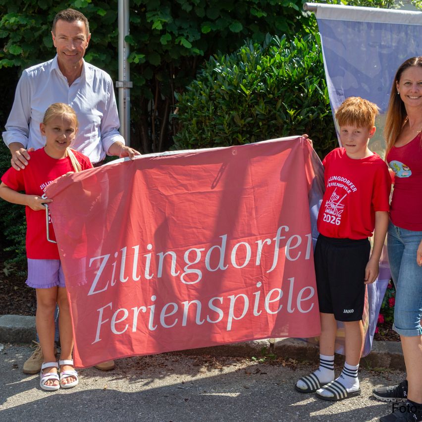 Ein Mann und zwei Kinder halten ein rotes Banner mit dem Text Zillingdorfer Ferienspiele und stehen neben einer Frau. Sie tragen alle rote Shirts und lächeln. Sie stehen auf einem Betonweg.