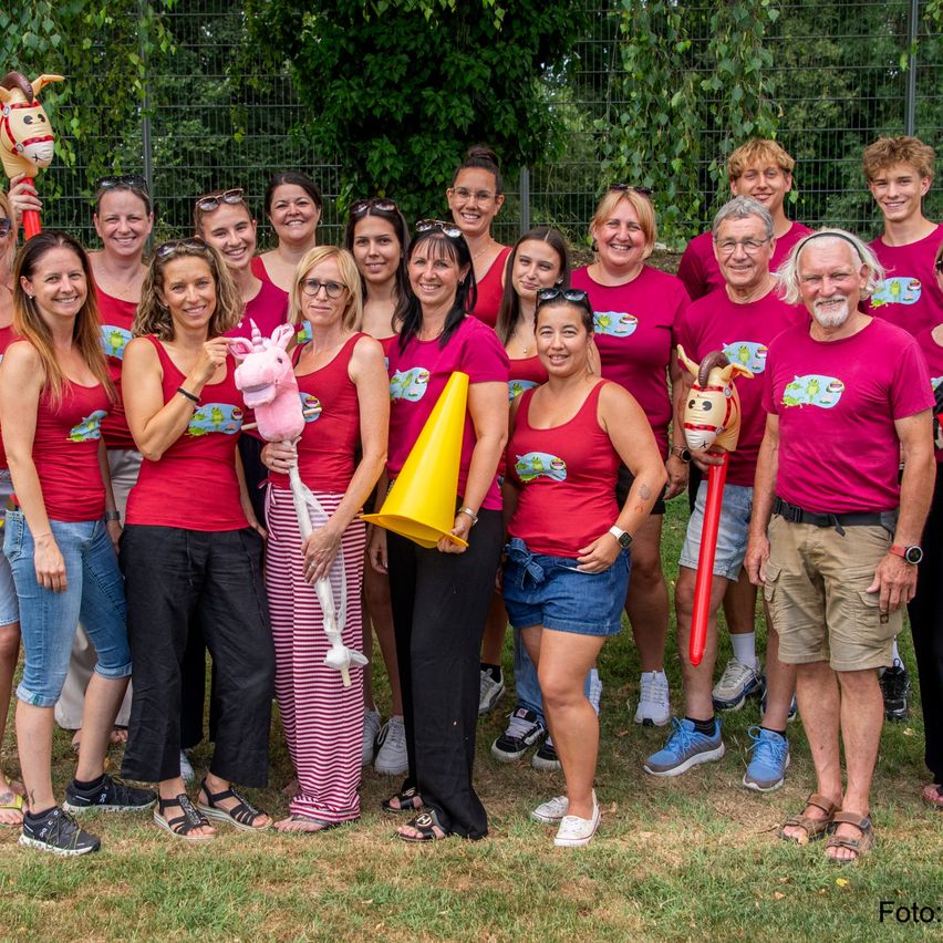 Gruppenfoto von Menschen in roten Shirts, die Plüschtiere, einen gelben Kegel und ein Pferdespielzeug halten. Sie stehen auf einer Rasenfläche.