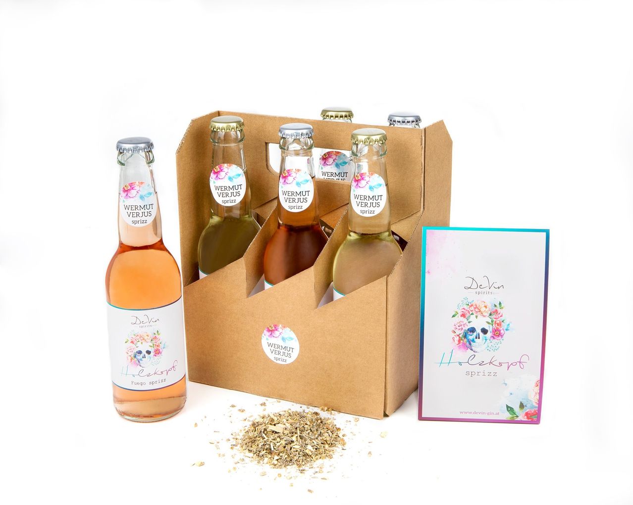 Eine Box mit sechs Flaschen Wermut Verjus Spritz, einem Kräutergetränk. Ein kleines Buch mit dem Titel Helkakopf spritz liegt neben der Box. Getrocknete Kräuter sind davor verstreut.