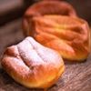 Zwei Gebäckstücke mit goldbrauner Kruste stehen auf einem Holztisch, bestreut mit Puderzucker. Das Gebäck auf der linken Seite hat eine leicht unebenmäßige Oberfläche, während das auf der rechten Seite glatter erscheint.