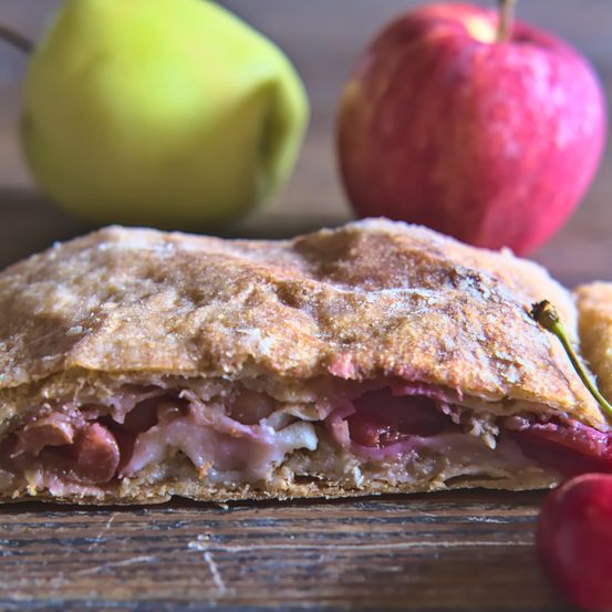 Ein Ausschnitt eines geschnittenen Kuchens mit Kruste auf einem Holztisch, begleitet von einer grünen Birne und einem roten Apfel.