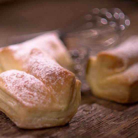 Nahaufnahme von zwei frisch zubereiteten Croissants, bestreut mit Puderzucker, auf einem Holztisch mit einem verschwommenen Schneebesen im Hintergrund.