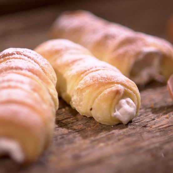 Drei Gebäckstücke, möglicherweise cremegefüllte Croissants, sind auf einer Holzoberfläche präsentiert. Sie sind mit Puderzucker bestreut und haben eine helle, goldbraune Farbe.