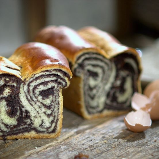Eine Nahaufnahme eines Brotes mit schokolade- und vanillefarbenen Marmorstreifen auf einer Holzoberfläche, mit einem zerbrochenen Eierschalenstück in der Nähe.
