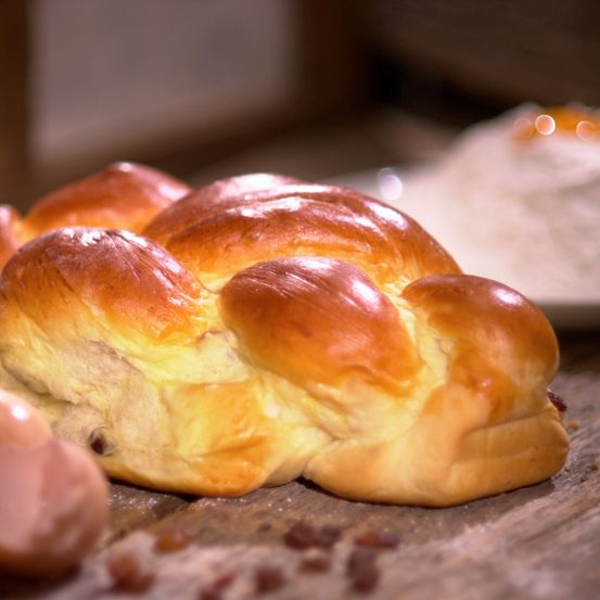 Ein Nahaufnahme eines gebackenen Challahbrots auf einem Holztisch, mit einigen Rosinen und Teig sichtbar.