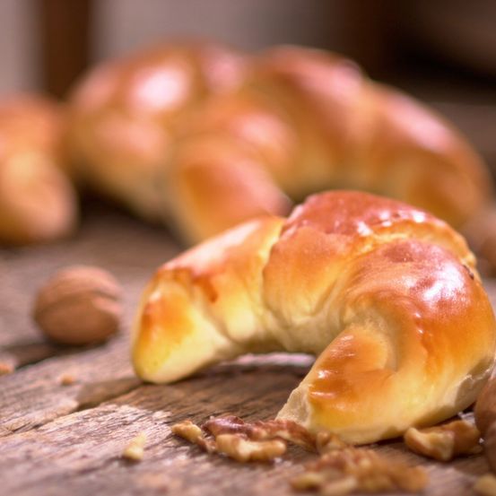 Ein Nahaufnahme eines goldbraunen Croissants auf einem Holztisch, mit ein paar umherliegenden Walnüssen. Das Croissant ist frisch gebacken, und seine Kruste ist leicht knusprig.