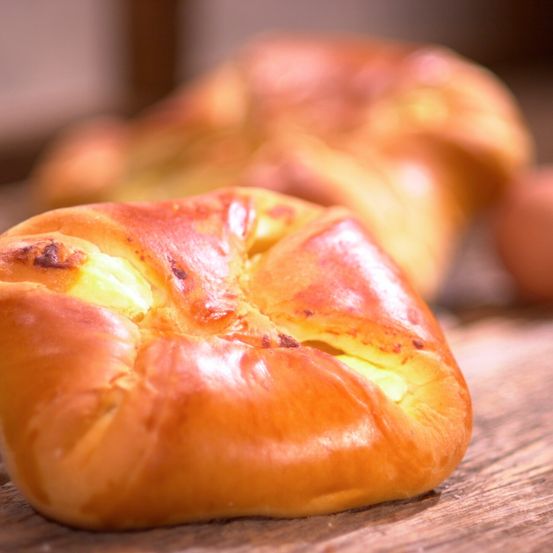 Eine Nahaufnahme eines goldbraunen Brötchens mit Käse, der aus der Mitte sickert, auf einer Holzoberfläche mit unscharfen Backwaren im Hintergrund.