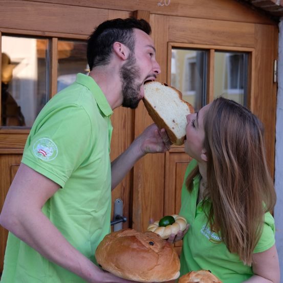 Ein Mann und eine Frau in grünen Uniformen stehen vor einer Holztür und teilen sich ein großes Brot, während der Mann ein Stück isst und die Frau das Brot küsst.