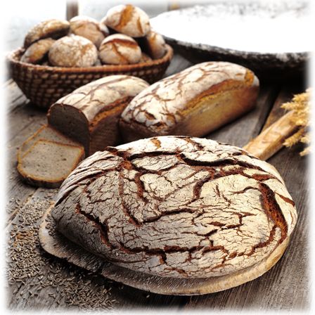 Auf einem Holztisch sind verschiedene Brote und Brötchen zu sehen. Zwei Brote sind ganz und eines ist geschnitten. Es gibt auch einen Korb mit Brötchen.