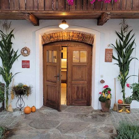 Ein rustikaler Holzeingang mit zwei Türen. Ein Schild sagt 'HOLZHAUS'. Dekoration umfasst Kürbisse und Pflanzen. Ein rotes Schild sagt 'Genuss Region'.