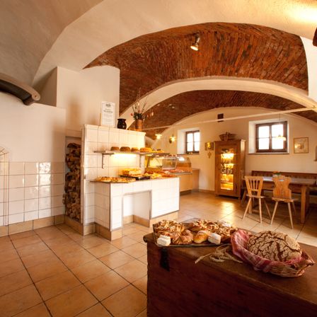 Ein rustikales Bäckerei-Interieur mit gewölbten Decken, einer Auslage mit Gebäck und einem hölzernen Tisch mit Stühlen. Holzregale und eine Theke mit mehr Backwaren.