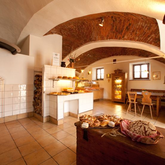 Ein rustikales Bäckerei-Interieur mit gewölbten Decken, einer Auslage mit Gebäck und einem hölzernen Tisch mit Stühlen. Holzregale und eine Theke mit mehr Backwaren.