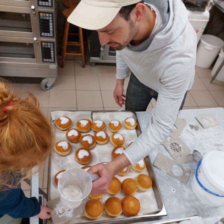 Ein Mann in einer weißen Kappe und einer grauen Kapuzenjacke gießt Puderzucker auf Donuts in einer Küche. Ein Mädchen mit roten Haaren beobachtet ihn von hinten.