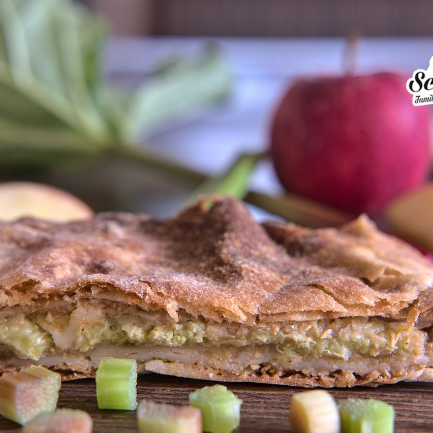 Ein Nahaufnahme eines mit Rhabarber gefüllten Kuchens, mit Rhabarberscheiben und einem Apfel im Hintergrund.