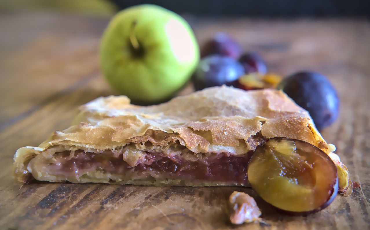 Ein Stück Obstkuchen mit Apfel- und Pflaumenfüllung, aufgeschnitten um die Schichten zu zeigen, auf einem Holztisch mit einem grünen Apfel und Pflaumen im Hintergrund.