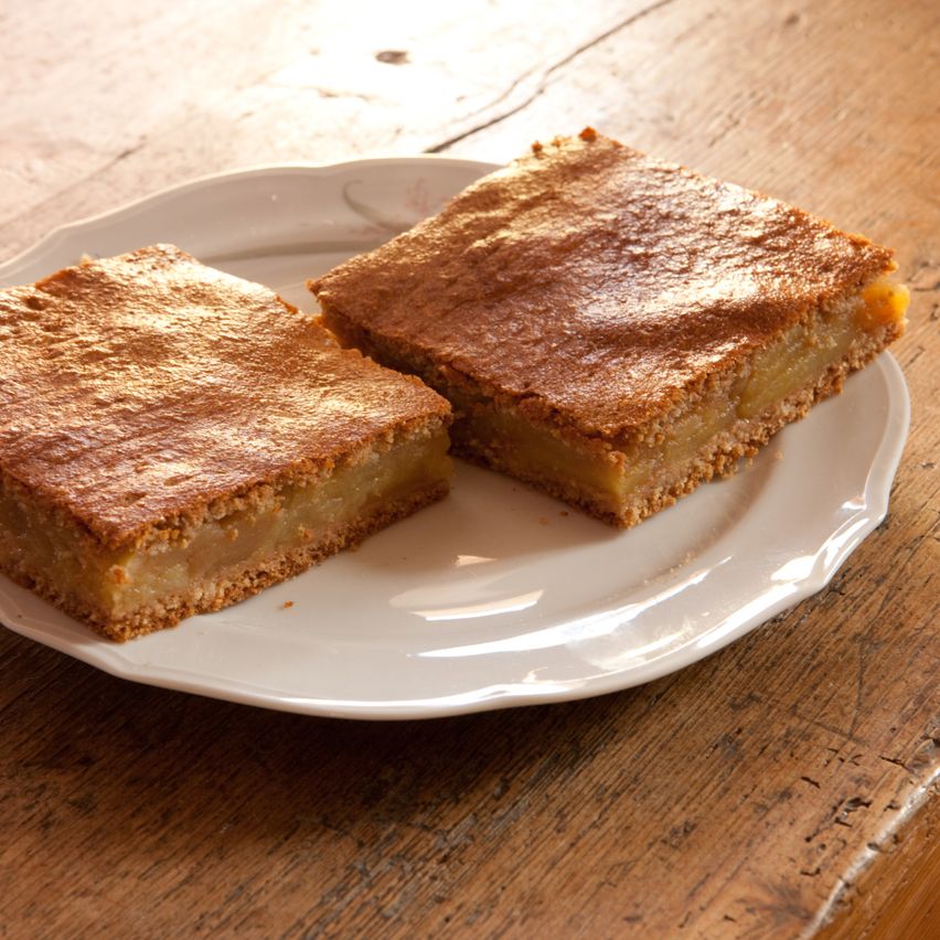 Zwei Stücke Apfelkuchen auf einem weißen Teller auf einem Holztisch. Der Kuchen hat eine goldbraune Kruste und ein sichtbares Apfelstück.