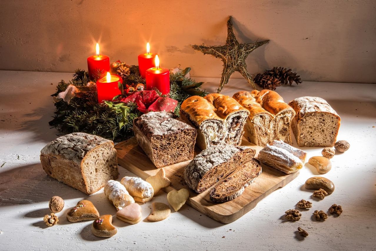 Eine Auswahl an Broten, Gebäck und Keksen liegt auf einem Holzbrett, begleitet von roten Kerzen, einem Kranz und einem Stern, was eine festliche Feiertagsatmosphäre schafft.