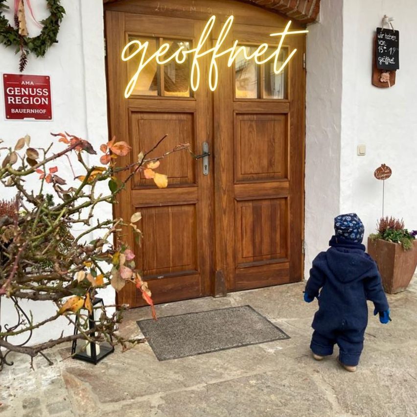 Ein Kind steht vor einer Holztür mit der Neonschrift 'geoffnet'. Die Tür wird von Pflanzen flankiert, über ihr befindet sich eine Tafel. Das Kind trägt eine blaue Jacke und Handschuhe.