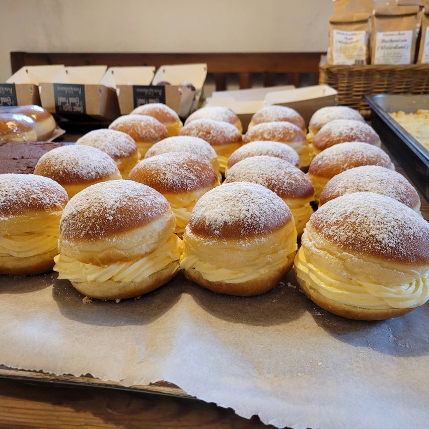 A close-up of a tray filled with several cream-filled donuts covered in powdered sugar, placed on a wooden table with more donuts and pastries in the background.