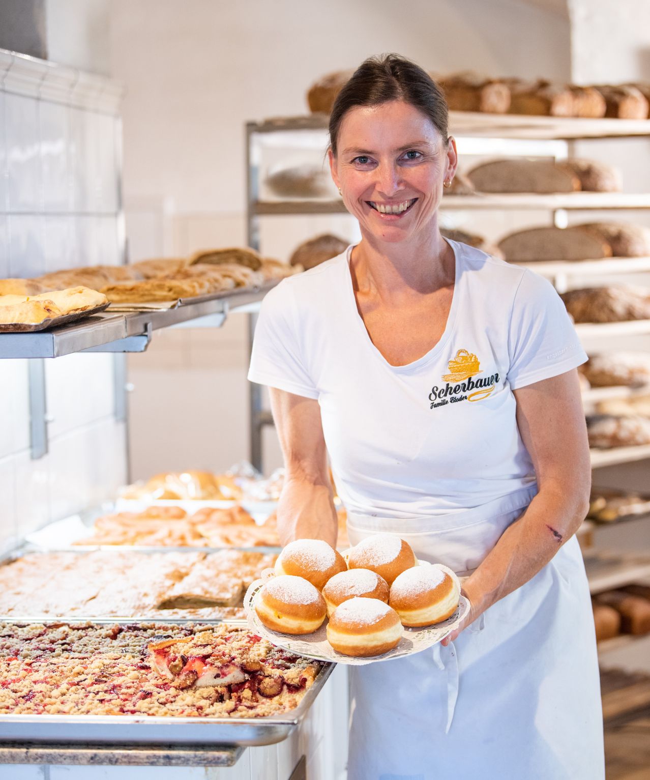A woman in a white apron stands in a bakery, smiling and holding a plate with donuts. Behind her, shelves are stocked with various baked goods.