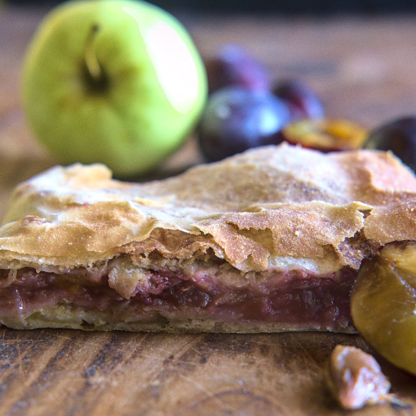 A close-up of a fruit pie slice, revealing a filling of plums and apples, on a wooden cutting board. A green apple is in the background.