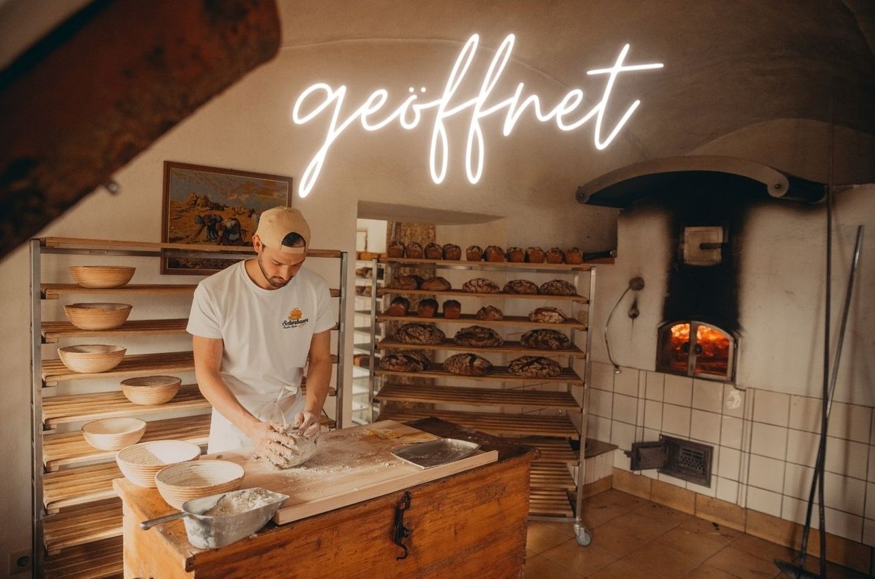 A man in a bakery prepares dough on a wooden board, with shelves of bread behind him and a neon sign above reading geoffnet.