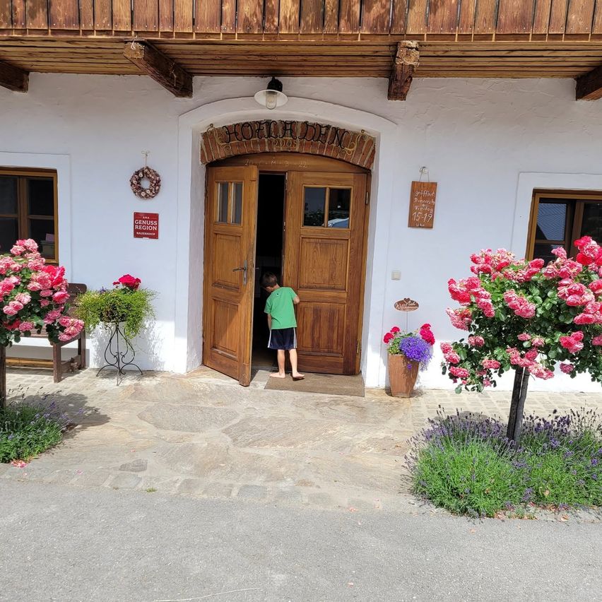 Ein junger Junge steht in der Tür eines traditionellen Holzgebäudes mit rosa Blumen und einem Kranz über dem Eingang.