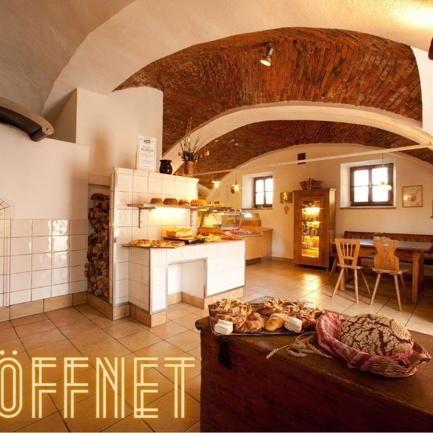 A cozy bakery with a wooden counter filled with bread and pastries, a wooden table with chairs, and a brick ceiling.