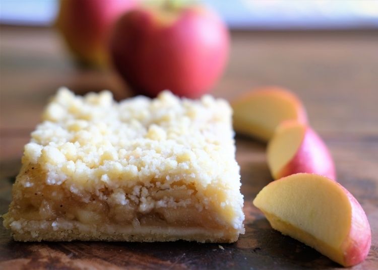 Ein Stück Apfelkuchen mit Streuseln steht auf einem braunen Holztisch. Dahinter sind Apfelscheiben, einige ganz. Der Tisch hat eine hellbraune Farbe.