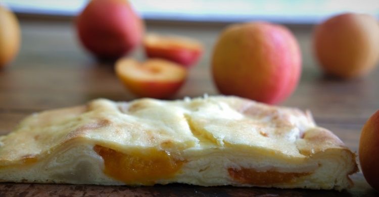 A slice of apricot tart sits on a wooden table with several whole apricots in the background, some cut and some whole.