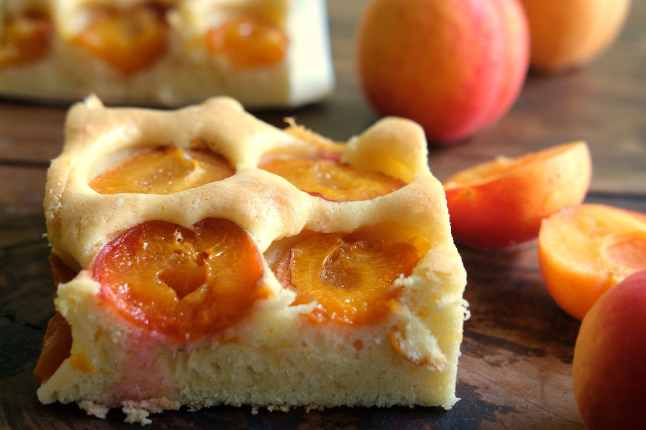 A slice of apricot tart shows its fruity filling and flaky crust. An entire tart and a few whole apricots are in the background.
