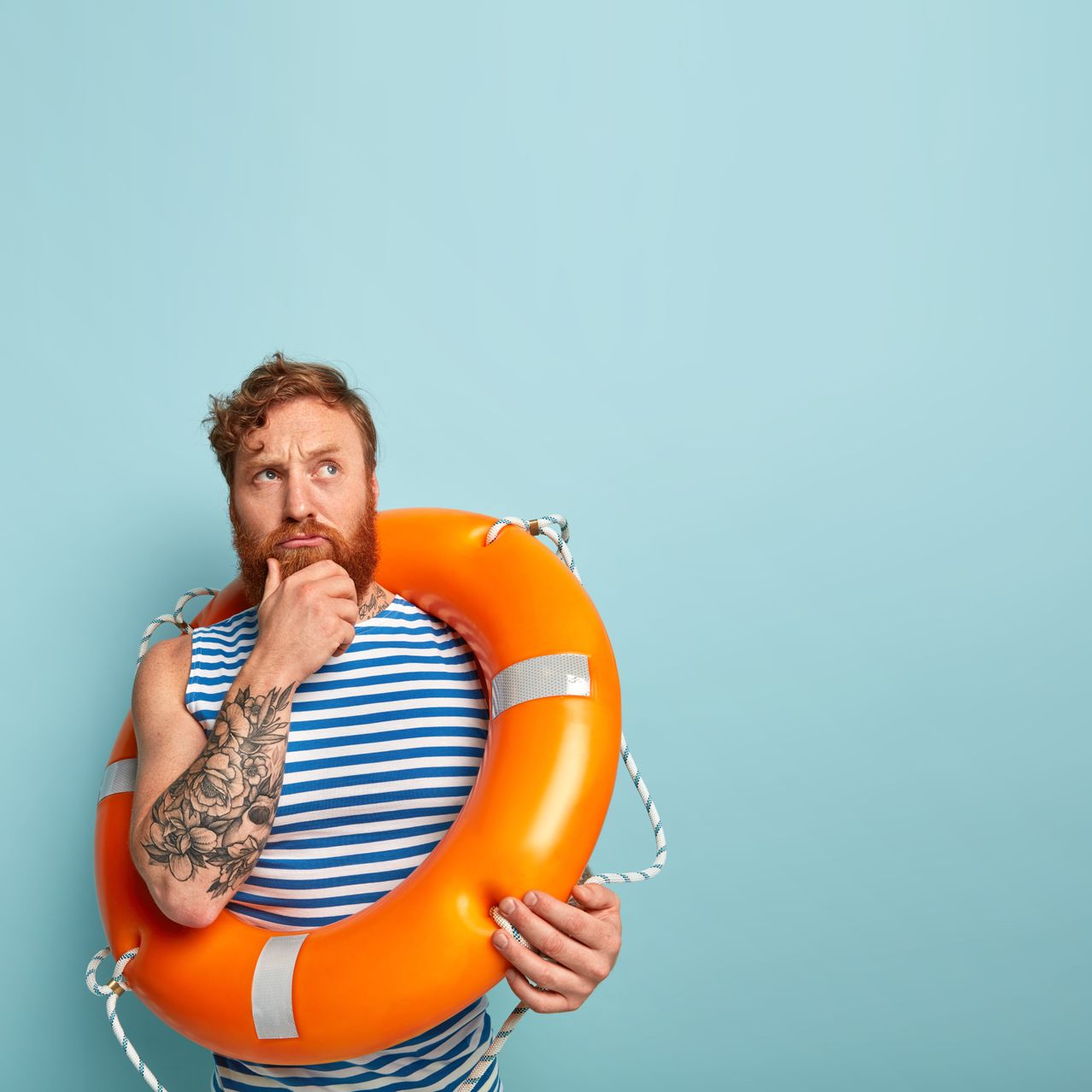 Ein bärtiger Mann in einem gestreiften Shirt hält einen orangefarbenen Rettungsring und steht vor einem blauen Hintergrund, er sieht nachdenklich aus.
