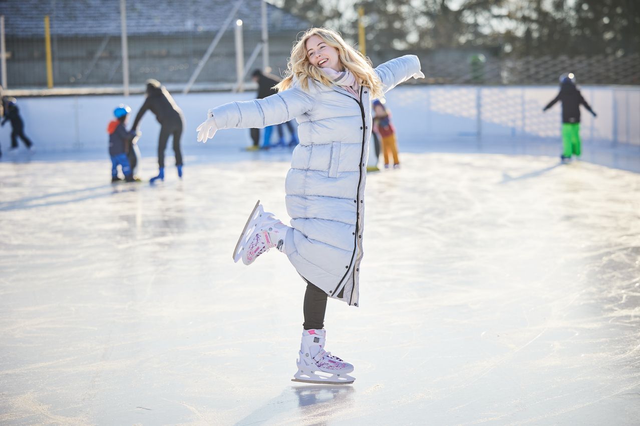 Eine Frau eisskatet auf einer Outdoor-Eisbahn. Sie hat ihre Arme weit geöffnet und lächelt. Hinter ihr eisskatende weitere Personen. Der Hintergrund ist unscharf.
