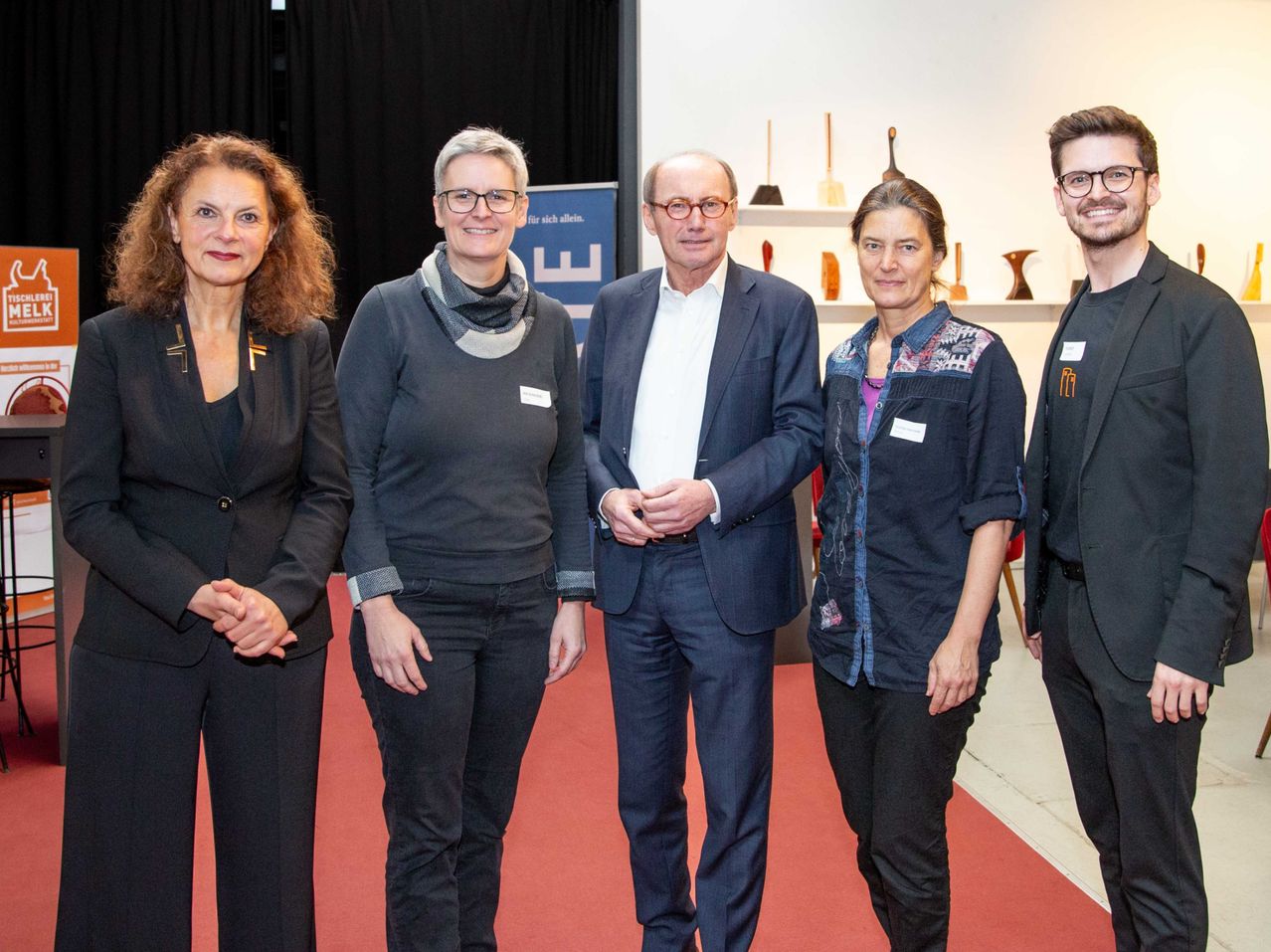 Fünf Personen stehen zusammen auf einem roten Teppich, wahrscheinlich bei einer Veranstaltung. Ein Mann in der Mitte trägt einen Anzug, während andere lässige Kleidung tragen. Sie scheinen für ein Foto zu posieren. Dahinter ist eine weiße Wand mit Regalen und einem Banner.