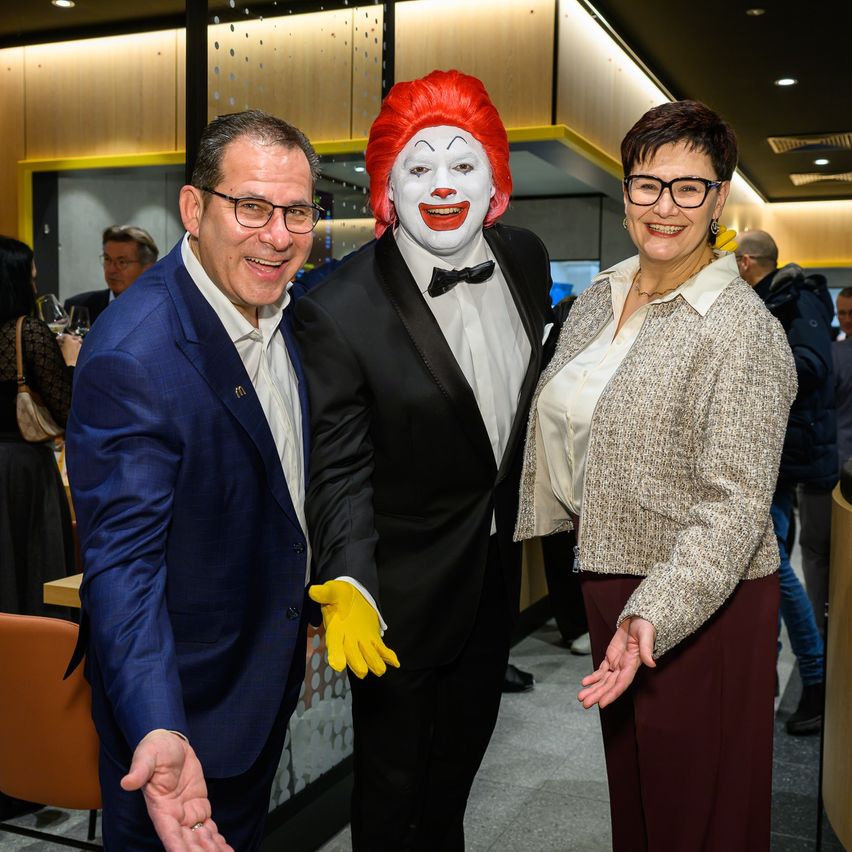 Drei Personen in einem Raum, zwei Männer und eine Frau, feierlich gekleidet, lächeln für ein Foto. Ein Mann ist als Ronald McDonald verkleidet.