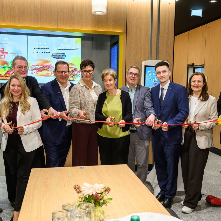 Eine Gruppe von Menschen in formeller Kleidung steht zusammen und schneidet ein Band vor einem Tisch mit Blumen. Dahinter zeigt eine große Leinwand eine Werbung für Burger.