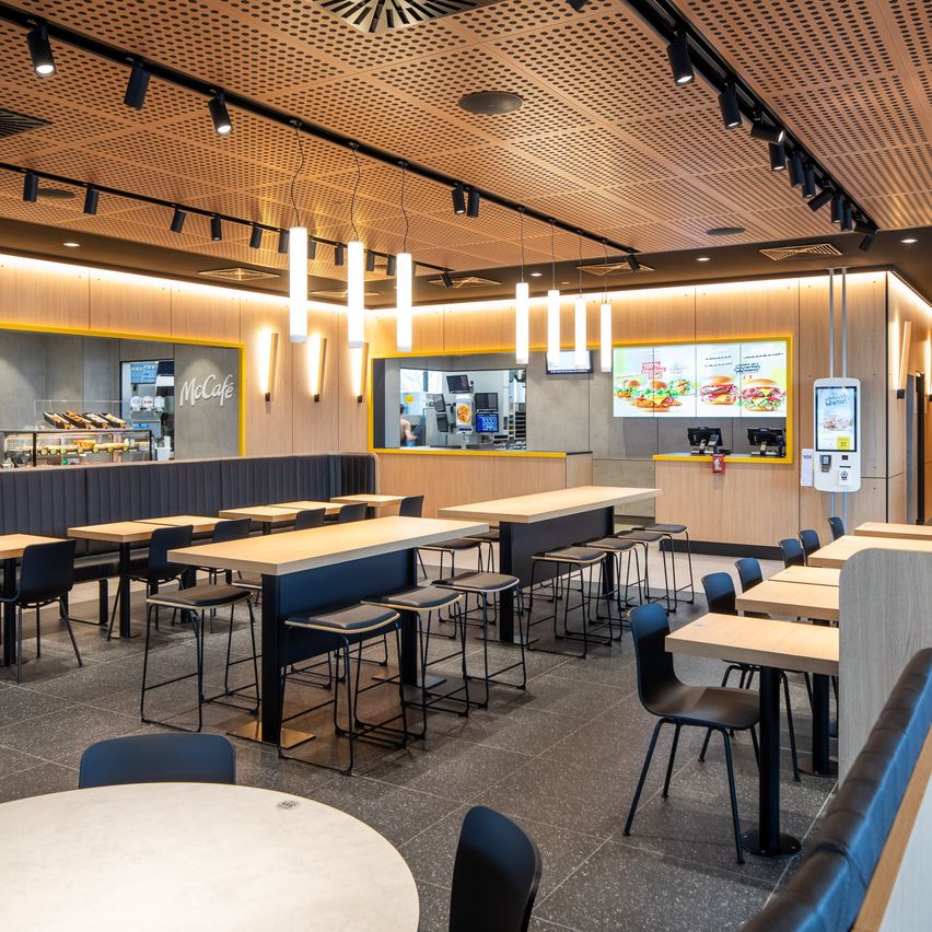 Innenansicht eines McDonald's-Restaurants mit modernen Möbeln, einer Theke mit Menütafel und Tischen mit Stühlen. Die Decke ist mit hängenden Lampen verziert.