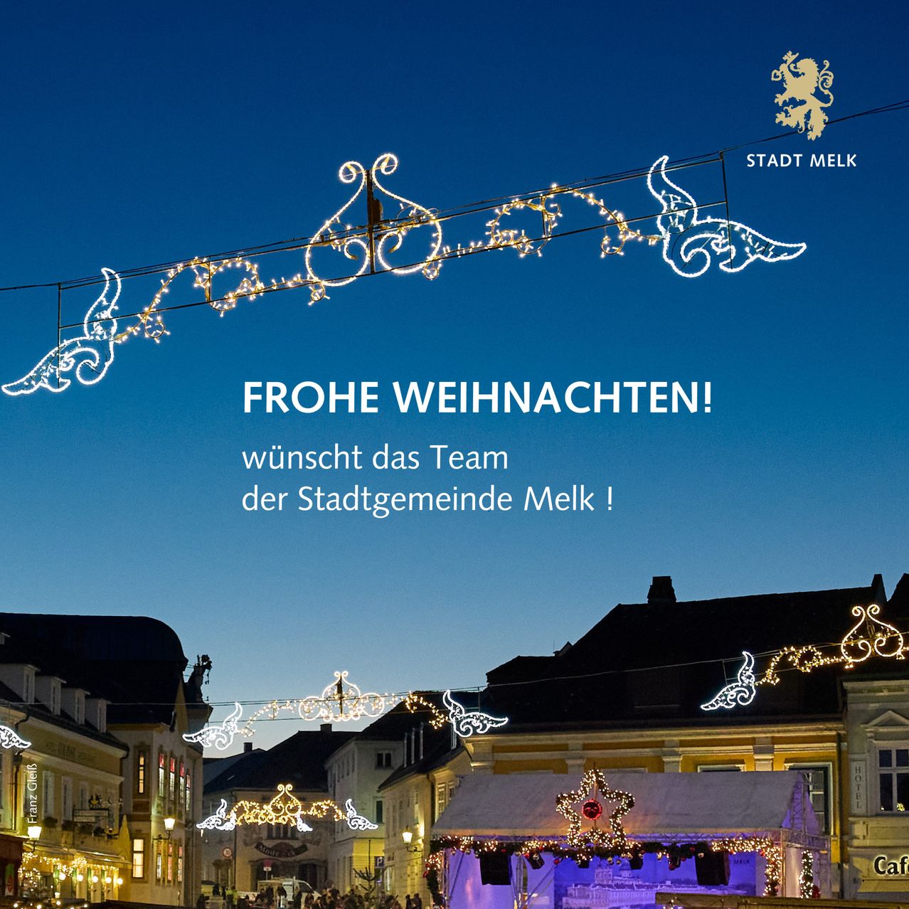 Ein abendlicher Stadtblick mit festlichen Lichtern und Dekorationen. Ein großes weißes Banner mit der Aufschrift 'Frohe Weihnachten!' Das Team der Stadt Melk wünscht Ihnen frohe Weihnachten.