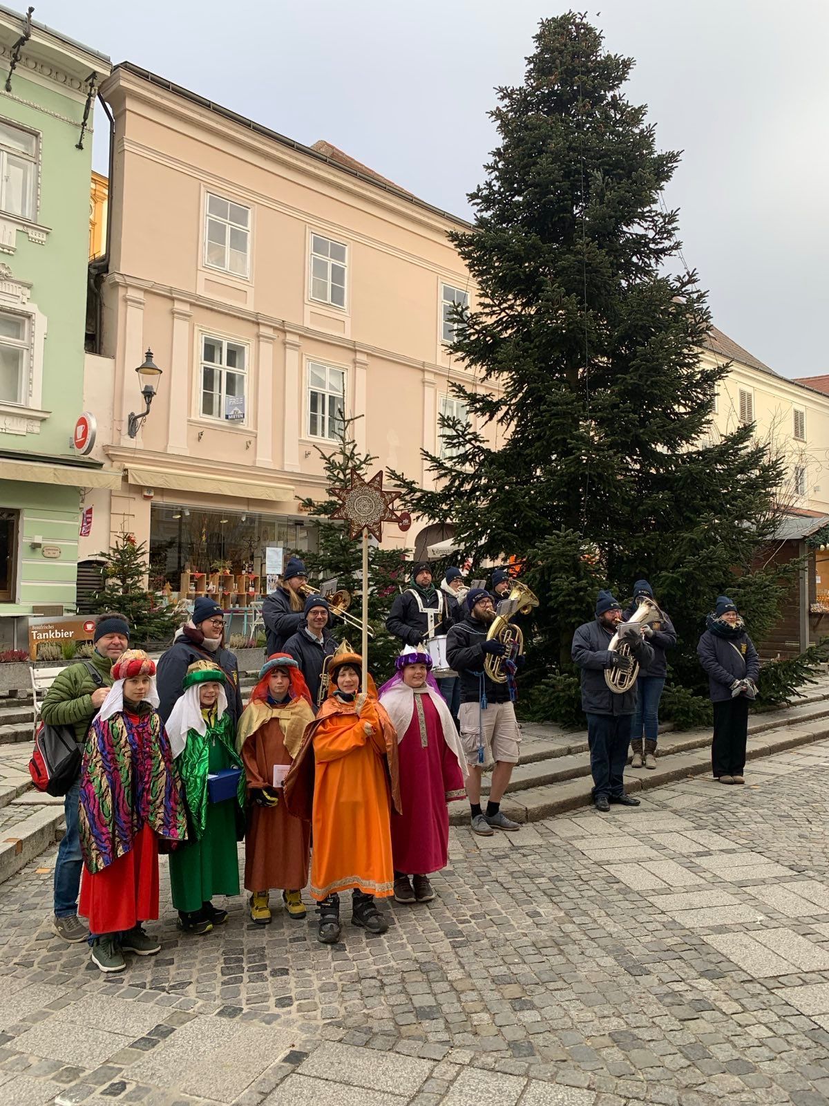 Eine Gruppe von Menschen in festlichen Kostümen posiert für ein Foto vor einem großen Weihnachtsbaum, während Musiker in der Nähe Instrumente spielen.