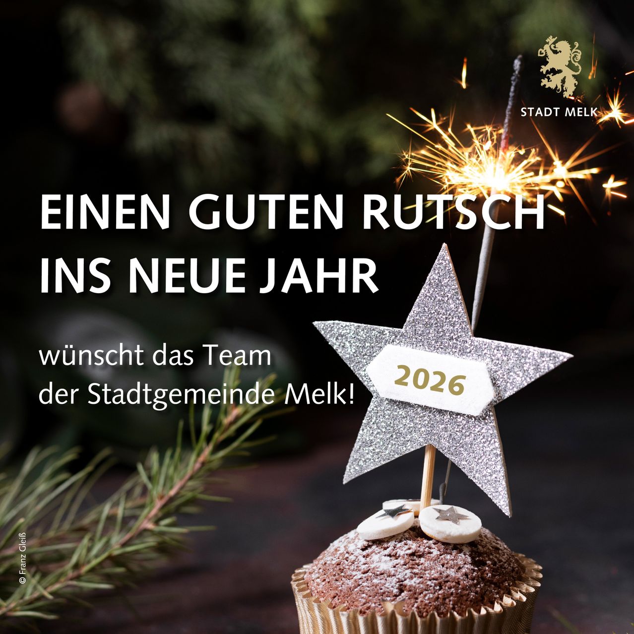 Ein Cupcake mit einem silbernen Stern oben und einem 2026-Schild ist von Feuerwerk umgeben. Er steht auf einer Holzoberfläche mit Tannenzweigen im Hintergrund. Der Text lautet 'Einen Guten Rutsch ins Neue Jahr'.