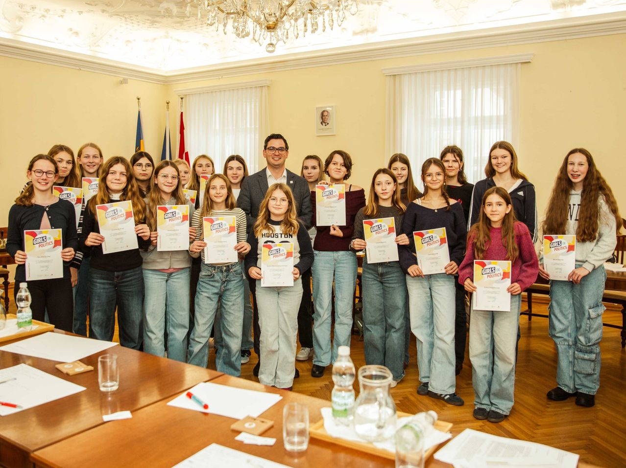 A group of girls and a man in formal wear stand in a room, holding certificates and smiling. They are in front of a desk with papers, glasses, and a bottle. Behind them, flags are displayed on poles.