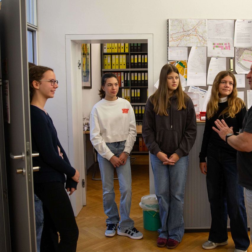 Five people are in a room with a wooden floor. Four women and one man. They are looking at something in front of them. Papers are pinned to the wall.