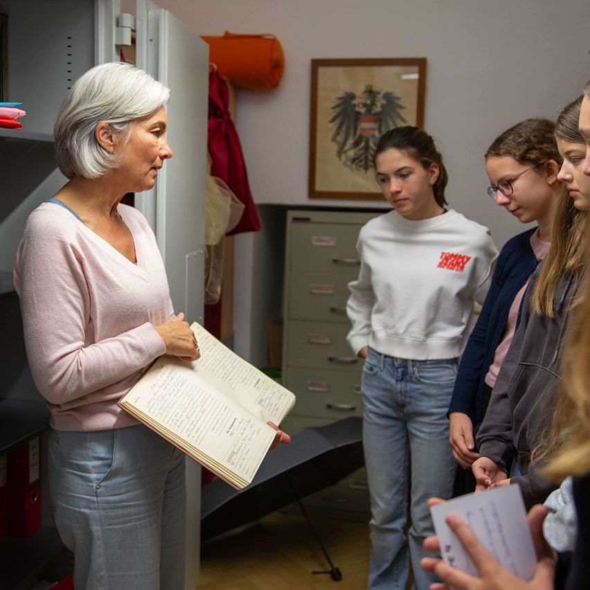 A woman holds an open book while looking at it. She is surrounded by four girls, one holding a piece of paper. They are in a room with a wooden cabinet and a framed picture on the wall.