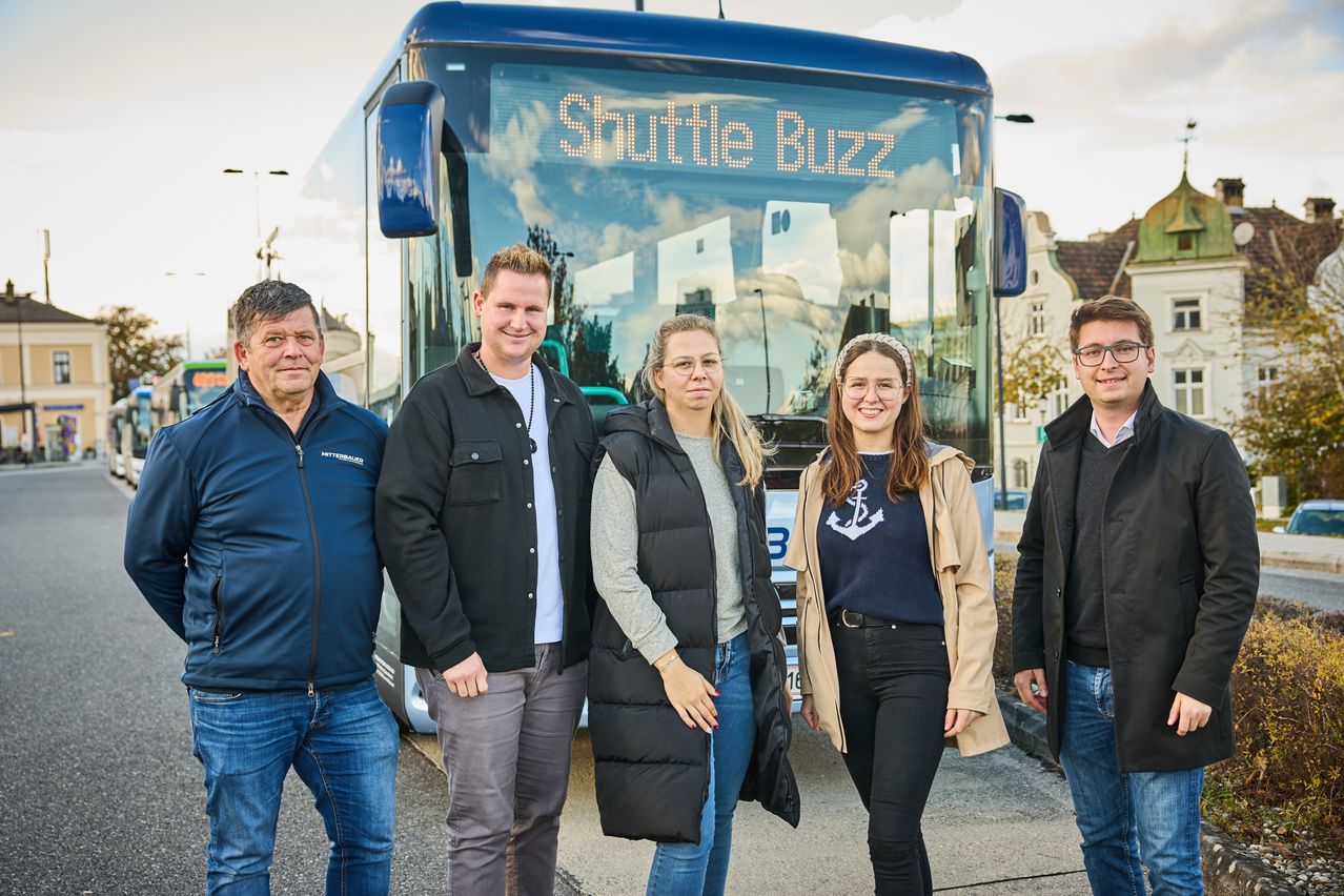 Fünf Personen stehen vor einem blauen Bus mit der Aufschrift 'Shuttle Buzz', der einen wolkenverhangenen Himmel im Fenster reflektiert.