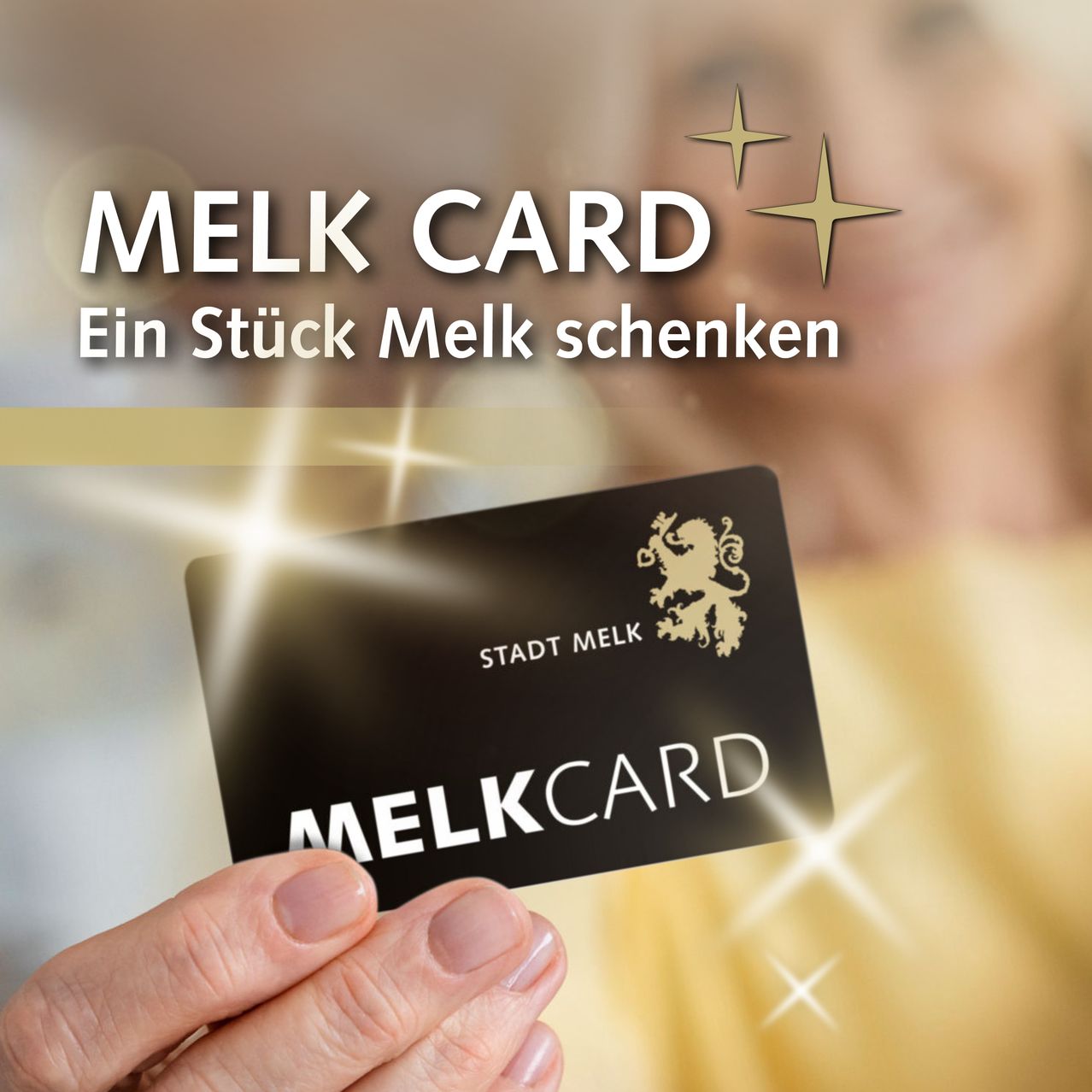 Eine Hand hält eine schwarze Karte mit der Aufschrift MELK CARD und einem goldenen Löwenlogo, vor einem verschwommenen Hintergrund mit Sternen und dem Text 'Ein Stück Melk schenken.'