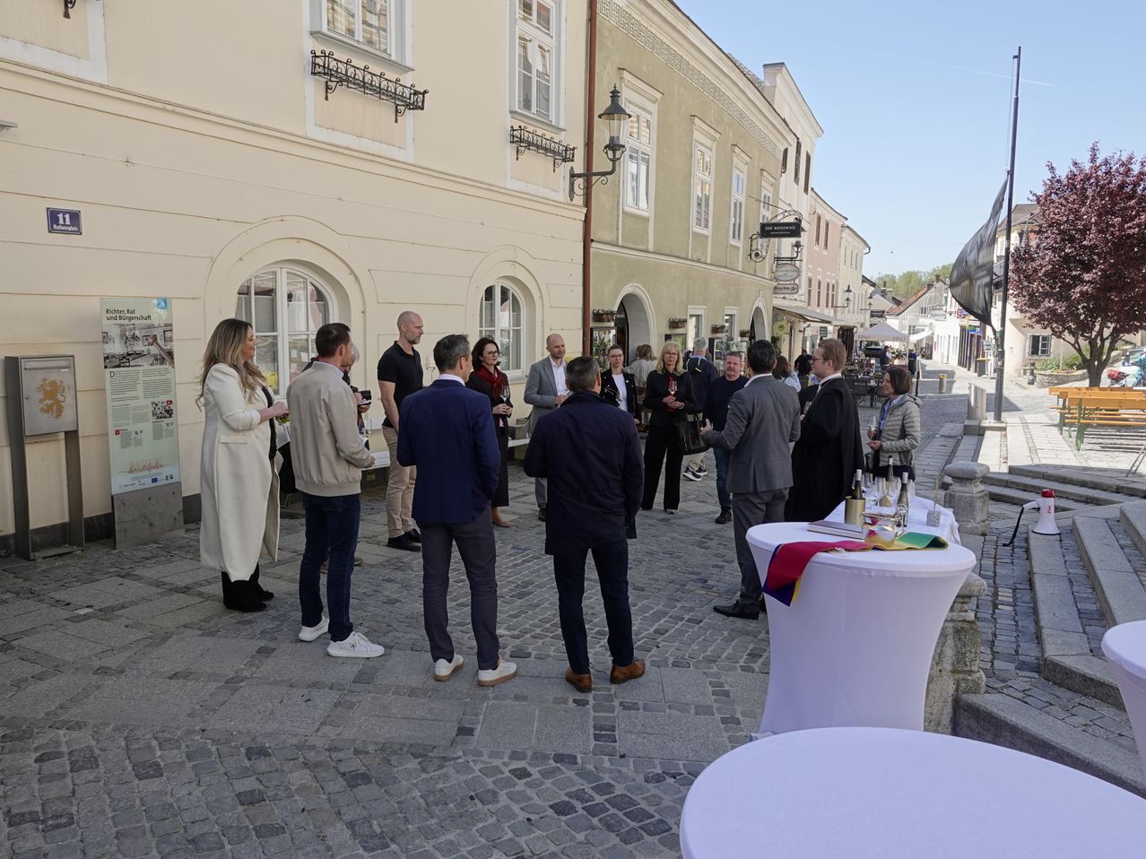 Eine Gruppe formell gekleideter Menschen versammelt sich auf einer Kopfsteinpflasterstraße, unterhält sich und netzwerkt möglicherweise. In der Nähe steht ein Tisch mit Weingläsern und anderen Gegenständen.