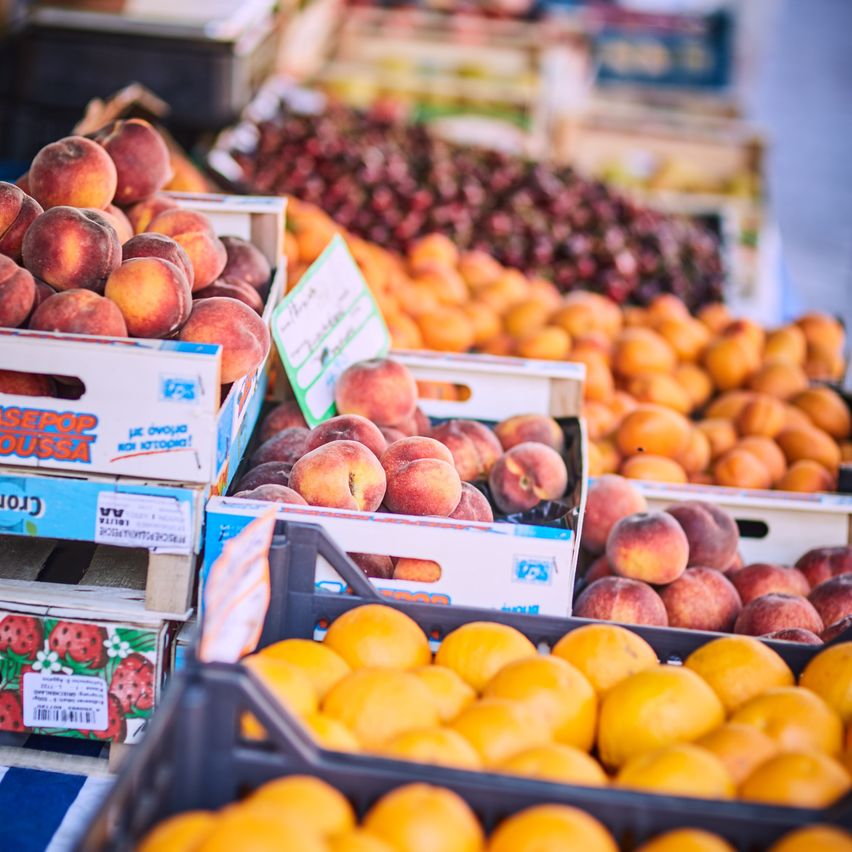 Mehrere Kisten mit Pfirsichen und Orangen sind auf einem Markttisch gestapelt. Die Pfirsiche sind in blauen und weißen Kisten, während die Orangen in schwarzen Körben sind.