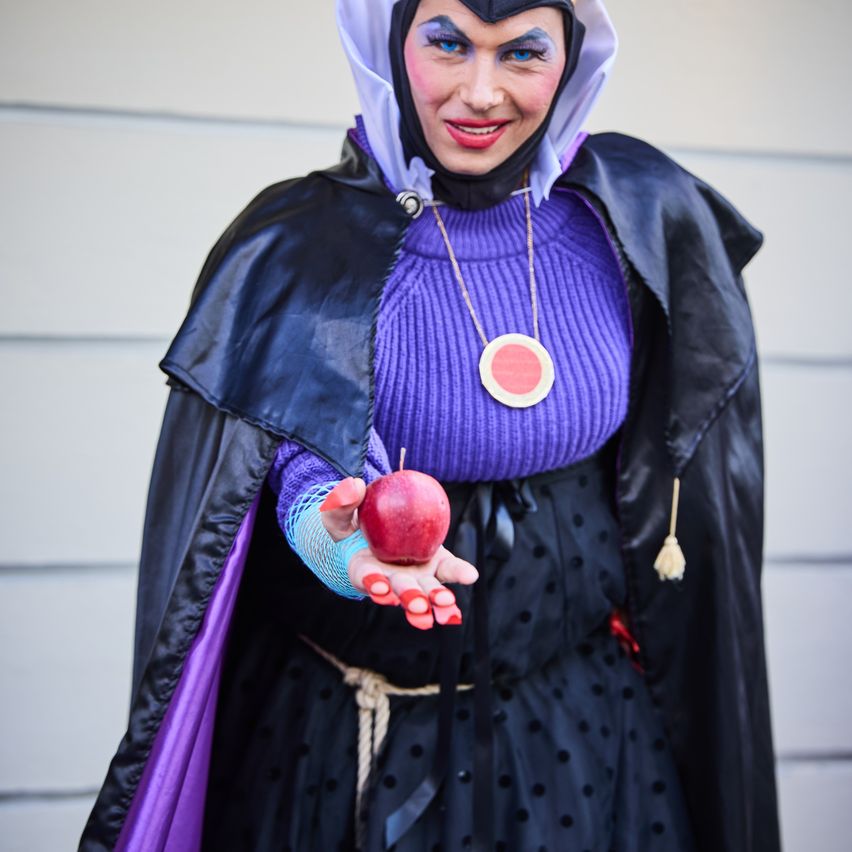 Eine Frau verkleidet als Malefiz aus Disneys Dornröschen. Sie hält einen roten Apfel. Sie trägt ein lila Oberteil, einen schwarzen Umhang und eine weiße Kapuze.