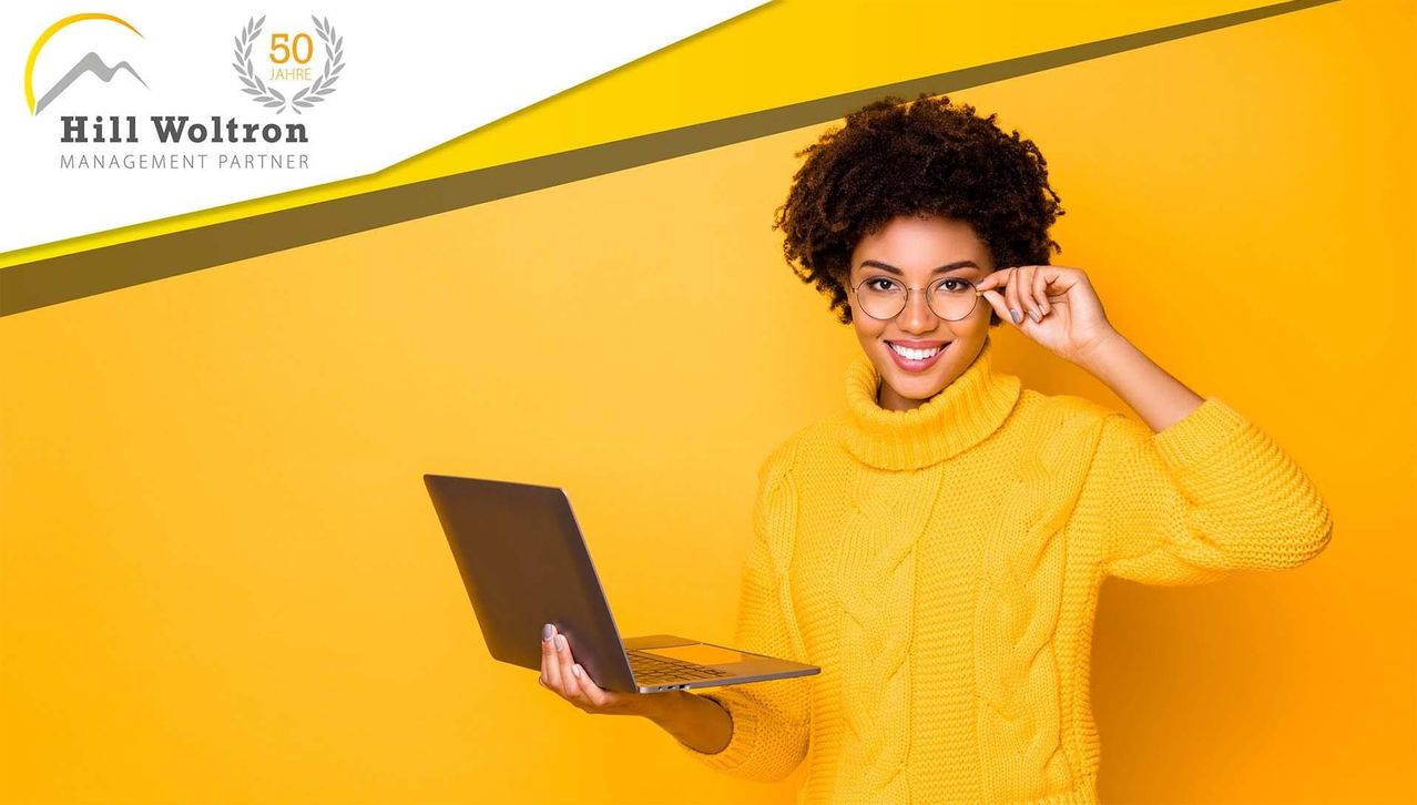 A woman with curly hair wearing a yellow turtleneck sweater and glasses holds a silver laptop in front of a yellow background.